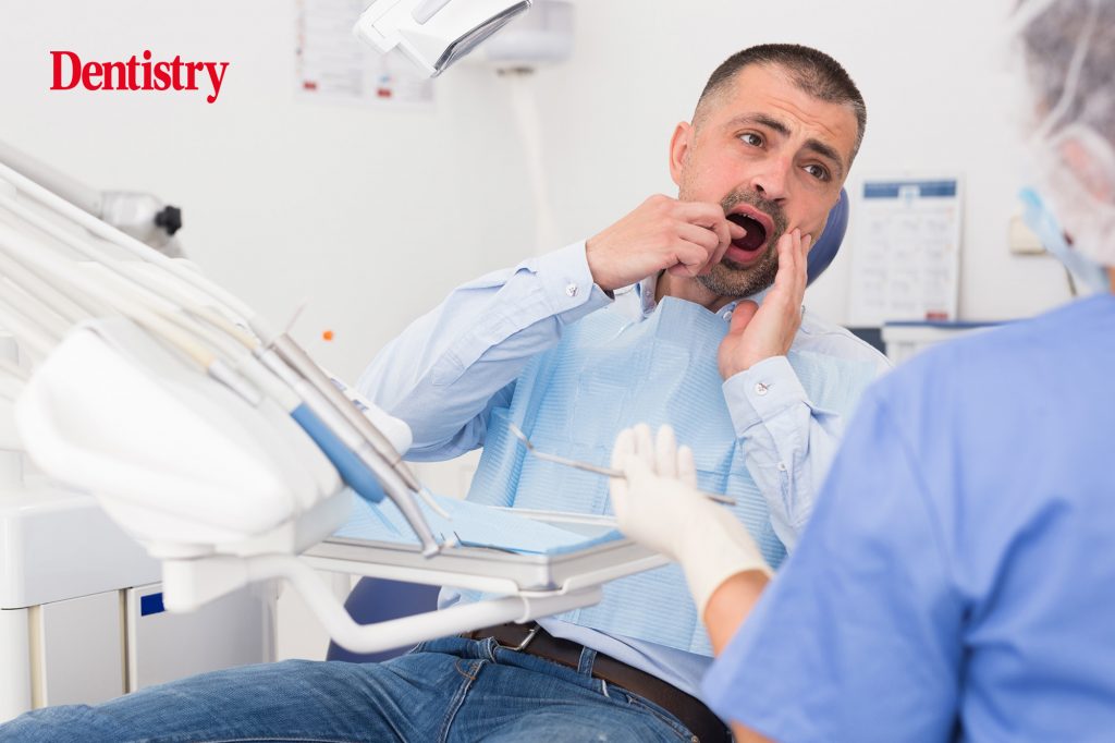 Unhappy patient pointing to his mouth in the dental chair while a dental nurse looks on